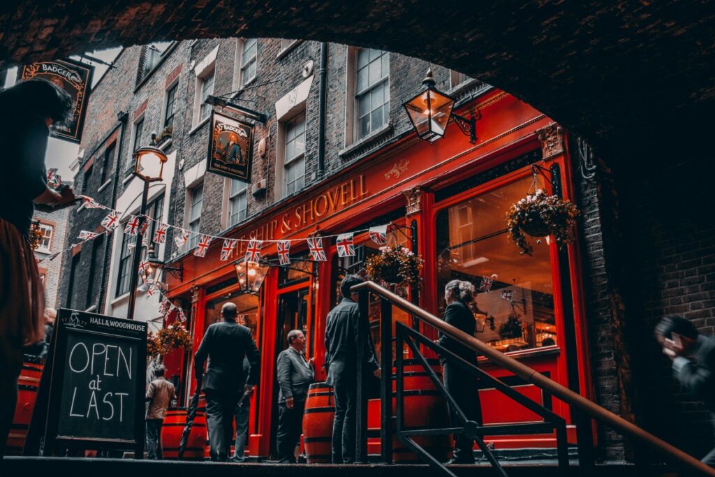 A charming red - fronted pub named "The Ship & Shovell" with British flags strung across, lanterns, and hanging flower baskets. People are gathered outside, and a sign reads "OPEN at LAST". 2025 UK Social Media Marketing Outlook: This inviting and traditional British scene could inspire social media marketing campaigns in 2025, highlighting the country's cultural allure.