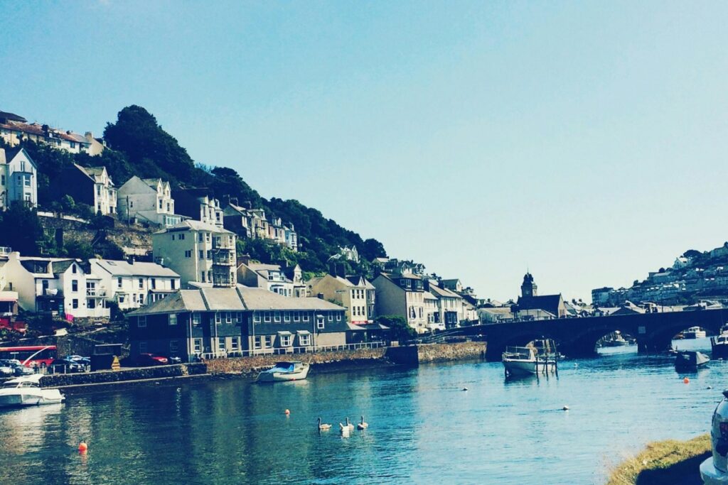 This image depicts a charming riverside scene in the UK. There are rows of houses built on a gentle slope, with a mix of white, beige, and grey exteriors, nestled among lush greenery. A bridge spans across the calm river, under which several boats are moored, and a few swans gracefully float on the water. To go viral on TikTok UK and boost your business, you can utilize images like this. Create engaging videos that showcase the idyllic British scenery, perhaps telling a story about a day in this riverside town, or highlighting local businesses around the area. Younger audiences in the UK love content that's visually appealing and tells an interesting story. Incorporating trends like challenges or using popular sounds while featuring such beautiful landscapes can increase your chances of going viral and driving more traffic to your business.