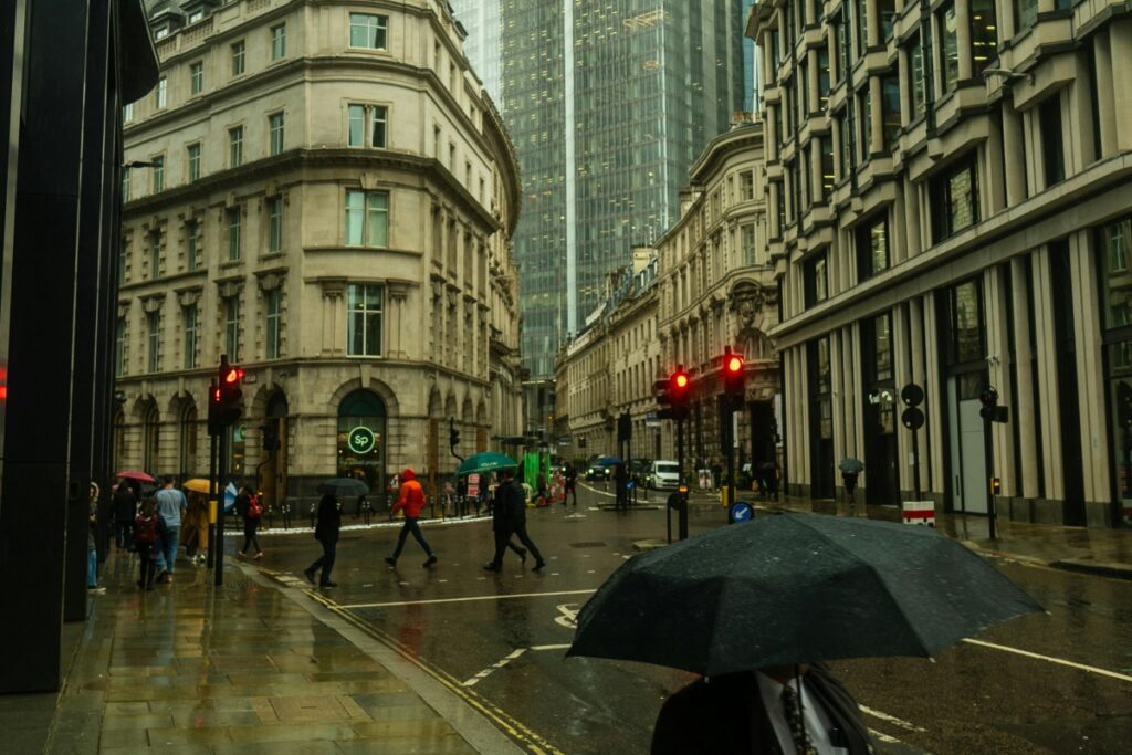 This image shows a rainy street scene in an urban area of the UK. Old - fashioned stone buildings line the street, contrasting with a modern glass skyscraper in the background. People with umbrellas are crossing the road at a junction, where traffic lights glow red. On Instagram UK, to grow your brand successfully, use images like this to tell a story. You can showcase the blend of tradition and modernity in the UK, which might resonate with your target audience. For instance, if your brand is related to fashion or lifestyle, you could talk about how your products fit into the dynamic urban life here. Engage with followers by asking questions about their experiences in such settings, and use relevant hashtags like #UKurbanlife, #Londonstreets (if applicable) to increase your reach and build a strong brand presence.
