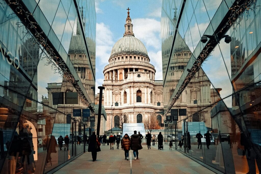 This image shows a bustling urban scene with people walking along a pathway flanked by modern glass - fronted buildings. At the end of the path stands the iconic St Paul's Cathedral, with its large dome and classical architecture. The glass buildings reflect the cathedral, creating an interesting interplay between the old and the new.